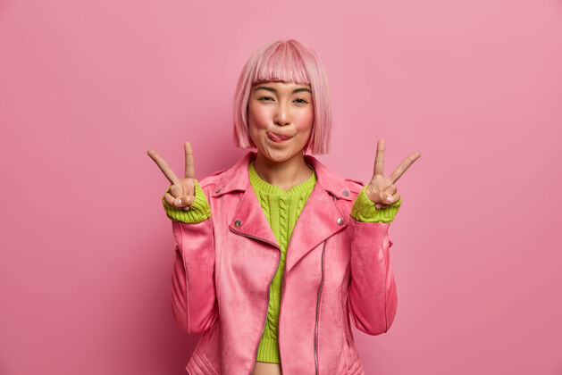 手指积极成功的亚洲粉红头发女人舔嘴唇 做胜利手势 露出两个手指 穿着时尚夹克 在室内摆姿势发型时尚热情