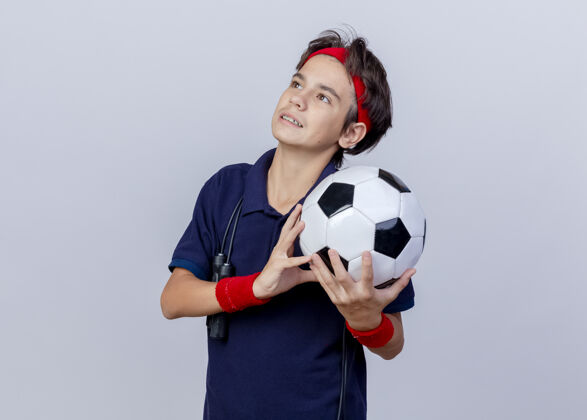 向上高兴的年轻帅气的运动男孩戴着头带和带牙套的腕带 脖子上系着跳绳 手里拿着足球 在白色背景上看着孤立的复制空间周围运动腕带