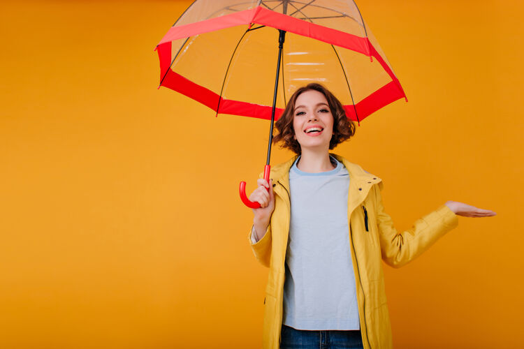 成人心情不错的好女孩 一边笑着 一边打着红伞摆姿势室内照片 时髦的高加索女士 带着闪亮的妆容 享受着阳伞的合影季节室内雨
