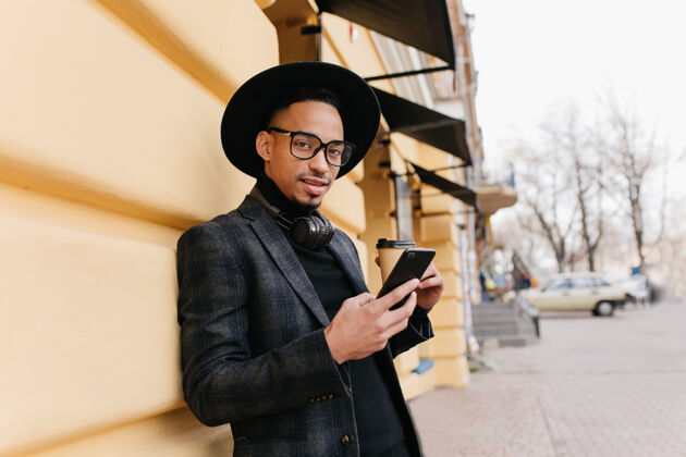 休闲可爱的棕色皮肤的年轻人站在老建筑附近 端着一杯咖啡时尚的非洲男模穿着休闲装 在街上摆着姿势 手里拿着电话电话手机男人
