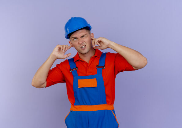 制服未出租的年轻男性建筑工人穿着制服 戴着安全帽 闭着耳朵站在紫色地上耳朵不愉快紫色