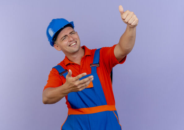 微笑微笑着的年轻男性建筑工人穿着制服 戴着安全帽 大拇指朝上放在紫色的地板上年轻紫色头盔
