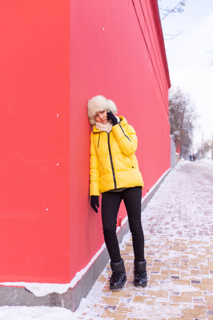 乐趣在一个冬天阳光明媚的日子里 一个穿着暖和衣服的红墙背景上的快乐的年轻女子在雪城的人行道上微笑着打电话说话手机科技