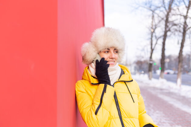 通话在一个冬天阳光明媚的日子里 一个穿着暖和衣服的红墙背景上的快乐的年轻女子在雪城的人行道上微笑着打电话手机电话漂亮