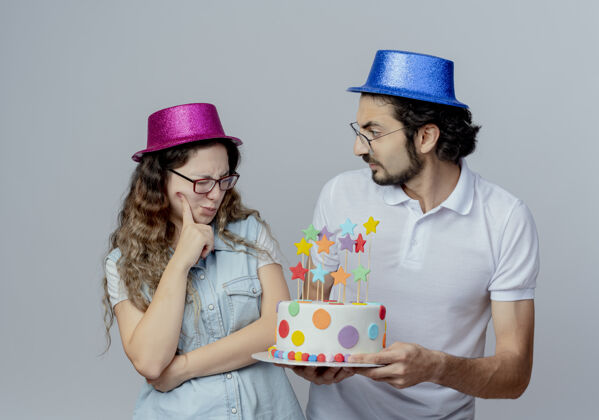 困惑小两口戴着粉蓝相间的帽子 小伙儿把生日蛋糕送给迷茫的女孩 孤立在白色的地板上年轻帽子生日
