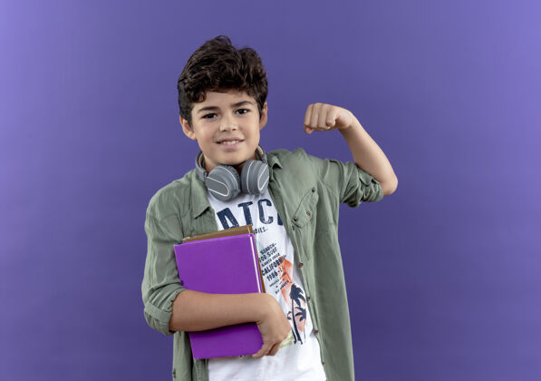小学生高兴的小男生戴着耳机拿着书 做着坚强的手势孤立在紫色的墙上手势请戴上墙