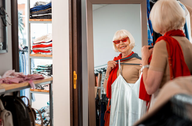 商品可爱的外表令人高兴的时尚的灰色头发的女人透过红色太阳镜看着购物镜 同时保持夏装在她面前反思老年人商业