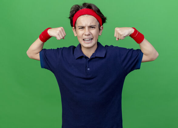 表情自信的年轻帅气的运动男孩戴着头带和护腕 戴着牙套 看着前面 做着一个强有力的手势 隔离在绿色的墙上背带佩戴腕带