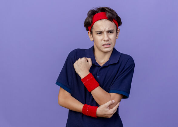 男孩年轻帅气的运动男孩戴着头带和护腕 戴着牙套 一边看着紫色墙上孤立的手肘表情壁板年轻