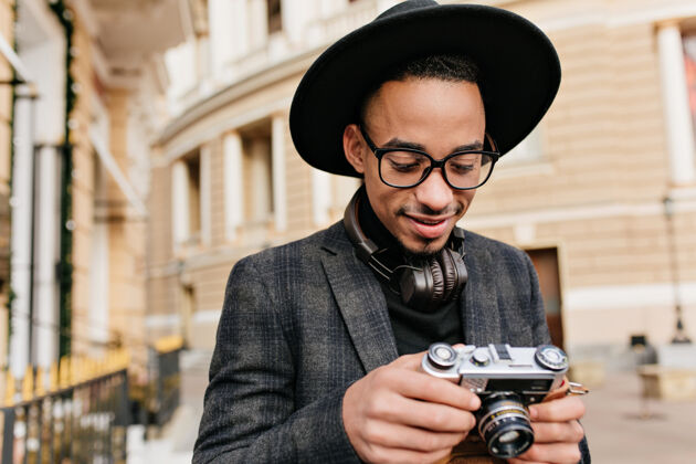 服装戴着耳机微笑的男摄影师站在城市街道上一张穿着格子夹克的快乐的非洲年轻人看着相机的户外照片时尚休闲服装