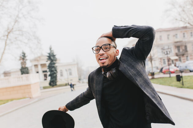 城市一个戴着大眼镜的非洲男人在公园里情绪激动地摆着姿势一个穿着格子灰色夹克的热情的黑人站在城市里休闲肖像城市景观