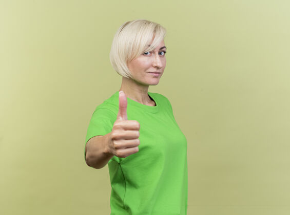 金发自信的中年金发女人站在侧视图上看前面 在橄榄绿的墙上孤立地竖起大拇指衣服感觉站着