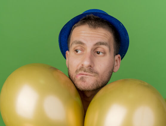 立场迷茫的帅哥戴着蓝色的派对帽站在氦气球旁边 看着隔离在绿色墙上的一面感情氦人