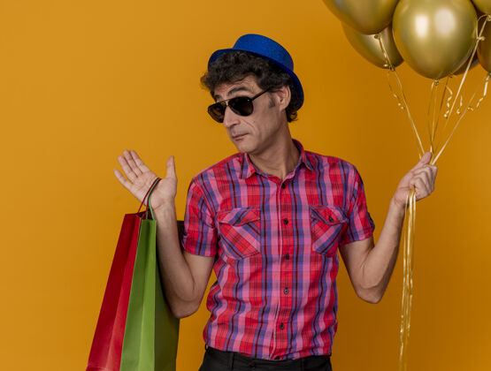 帽子笨手笨脚的中年人戴着聚会帽 戴着墨镜 手里拿着气球和纸袋 看着前面 一只空手孤立在橙色的墙上表演纸金