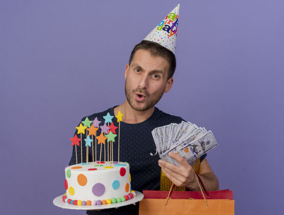 衣服印象深刻的帅哥戴着生日帽拿着生日蛋糕纸购物袋礼盒和钱隔离在紫色的墙上钱男人生日