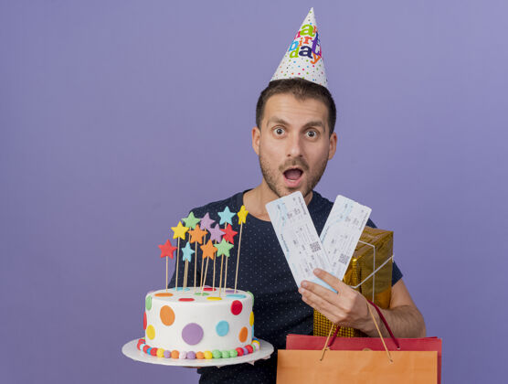 英俊兴奋的帅哥戴着生日帽拿着生日蛋糕纸购物袋礼盒和机票隔离在紫色的墙上蛋糕帽子表情