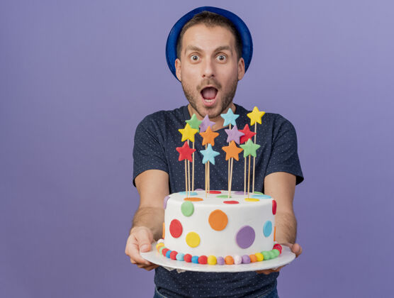 兴奋兴奋的帅哥戴着蓝色的帽子拿着生日蛋糕看着前面隔离的紫色墙壁男人衣服人