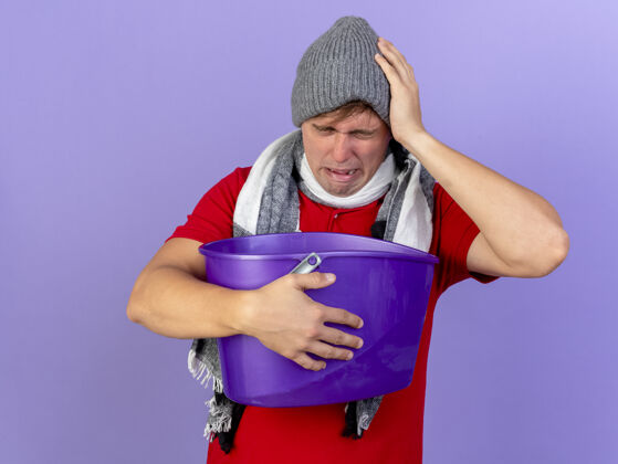 帽子疼痛的年轻英俊的金发病男子戴着冬天的帽子和围巾拿着塑料桶触摸隔离在紫色墙上的头表情围巾疼痛