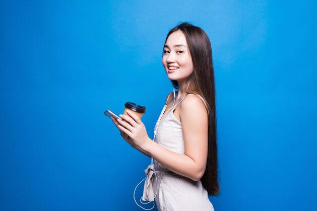咖啡照片中的亚洲年轻女子微笑着站着 用手机捧着咖啡纸杯 看着蓝色墙上的智能手机杯子女士女性