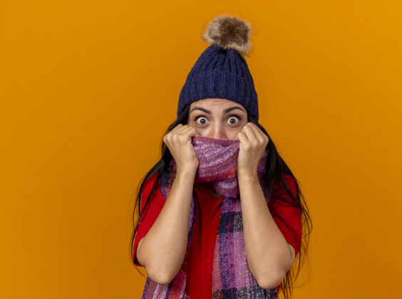 封面印象深刻的年轻生病的女人戴着冬天的帽子和围巾 看着前面用围巾遮住嘴巴隔离在橙色的墙上印象冬天女人