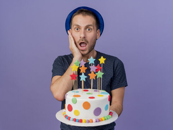 人兴奋的帅哥戴着蓝色的帽子把手放在脸上 拿着生日蛋糕孤立地放在紫色的墙上感觉衣服穿着