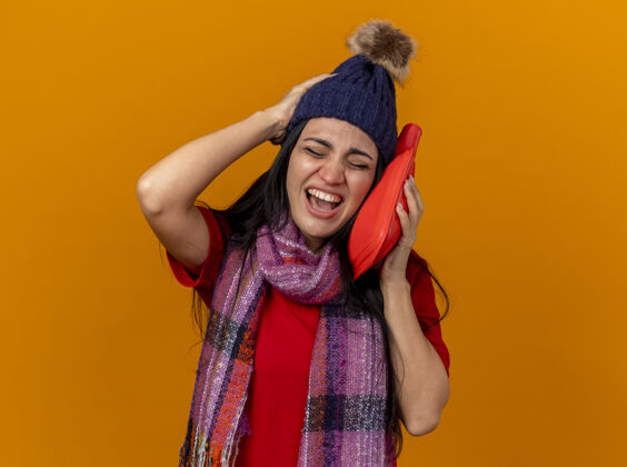 帽子带着冬天帽子和围巾的疼痛的年轻生病的女人用热水袋触摸着脸 手放在头上 闭着眼睛隔离在橙色的墙上站立包衣服