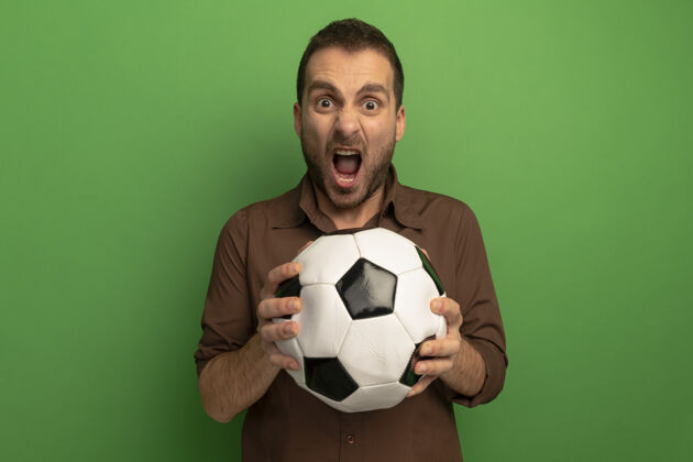 年轻人愤怒的年轻人拿着足球看在前面尖叫隔离在绿墙上人足球感觉