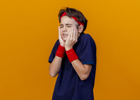 人疼痛的年轻英俊的运动男孩戴着头带和戴着牙套的腕带 手放在脸上 闭着眼睛孤立在橙色的墙上脸手穿着