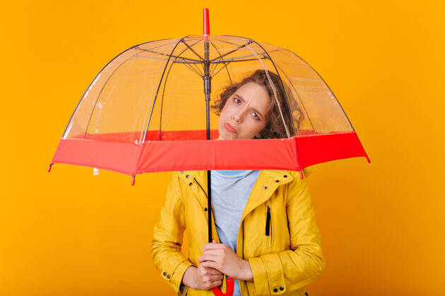 肖像一个心烦意乱的黑发女孩在雨伞下摆姿势一个穿着雨衣的悲伤的高加索女士在明亮的墙上拿着阳伞的画像年轻成人卷发