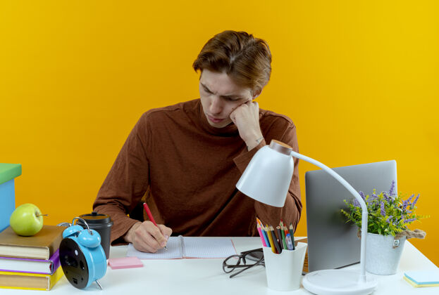 笔记本思考的年轻学生男孩坐在书桌旁 拿着学习工具在黄色的笔记本上写东西写作思考工具