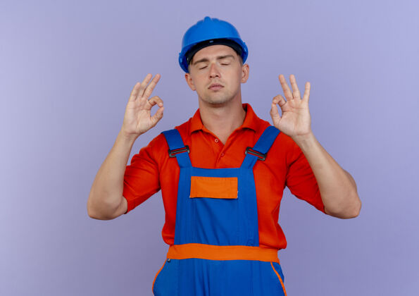 年轻年轻的男性建筑工人闭着眼睛 穿着制服 戴着安全帽 在紫色的地板上摆出一副很好的姿势眼睛头盔安全