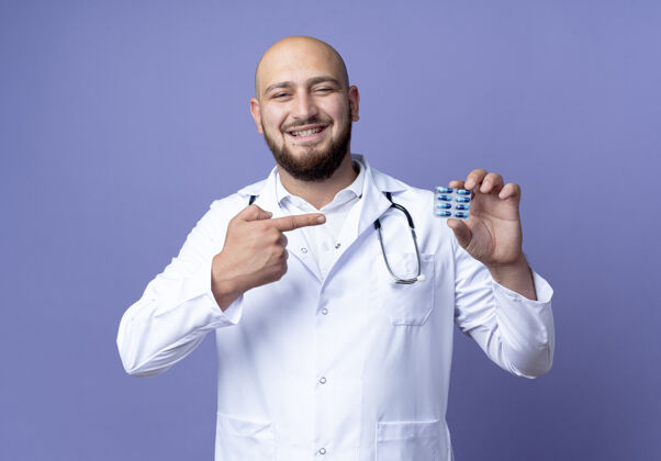 医生面带微笑的年轻秃头男医生穿着医用长袍和听诊器长袍药丸听诊器