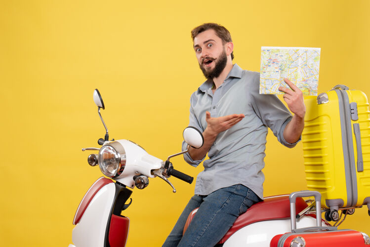 摩托车旅游概念的前视图 好奇的年轻人坐在摩托车上 手提箱放在摩托车上 指着后面 拿着黄色的地图地图手提箱拿着
