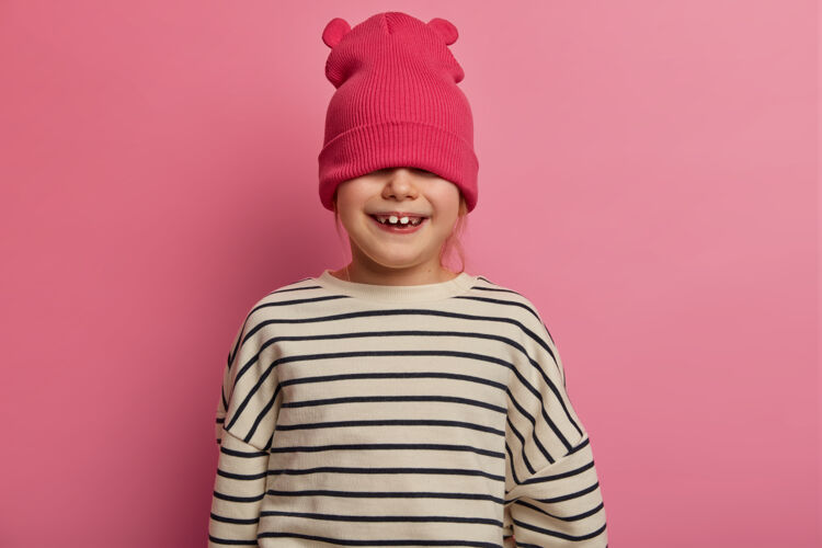 红发顽皮的小女孩戴着时髦的帽子 藏着眼睛 玩得很开心 不想去幼儿园 有着牙齿般的微笑 心情非常乐观 在柔和的玫瑰色墙壁上摆姿势孩子 时尚 时尚隐藏姜罗西