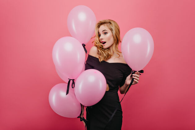 年轻惊讶的金发女人拿着漂亮的派对气球惊讶的可爱女孩穿着黑色的衣服孤立在粉红色的墙上女人服饰玩