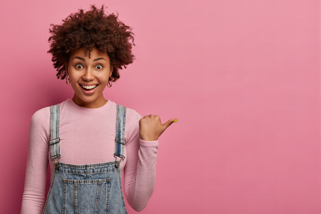 女孩积极的非裔美国女孩指着拇指 在空白的粉色墙上展示复制空间 有愉快友好的表情 穿着随意 在室内摆姿势 建议向右走 说沿着这个方向走促销卷发欣喜若狂