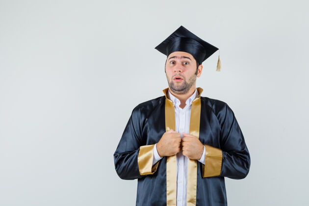 欢呼年轻人拿着拳头在他的毕业制服礼服 看起来困惑不解前视图朋友自信成功