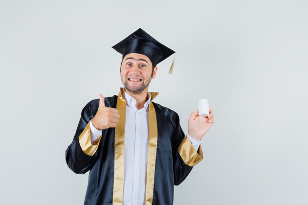 毕业身着毕业制服的年轻人手里拿着一瓶药丸 竖起大拇指 看上去很高兴 正对着前方拇指前面朋友