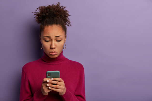 电话人 不快乐的情绪概念伤心失望的女人收到不好的短信 拿着智能手机 不满地集中在屏幕上 穿着紫红色圆领衫 在室内摆姿势 感到孤独人情绪悲观