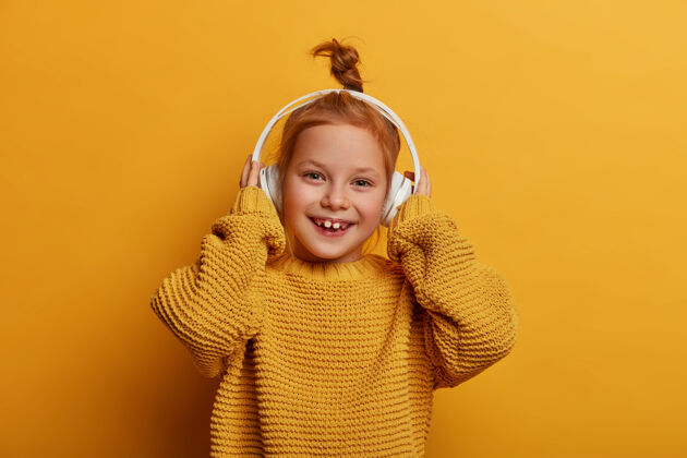 脸积极向上的快乐姜女通过耳机收听音轨 享受喜爱的爱好 穿着特大号针织毛衣 隔着黄色的墙壁孩子 音乐和乐趣的概念乐观小情绪