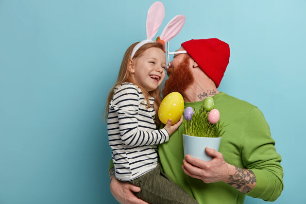 童年贴心的父亲亲吻女儿 牵手 捧着一盆彩蛋 准备复活节快乐的姜姑娘戴着兔子耳朵 背着大黄蛋宗教节日 庆祝理念男人家庭兔子