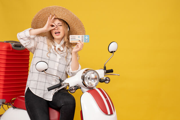 顶俯视图 微笑的年轻女子戴着帽子 坐在摩托车上 拿着制票眼镜 黄色的手势使坐年轻女子