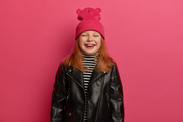装束时髦的姜可爱的女孩真诚地笑着 闭上眼睛 感到很高兴 欢庆美好的一天 和爸爸妈妈一起度过空闲时间 戴着粉红色的帽子和皮夹克 准备上学皮革高兴人