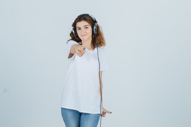 肖像身穿白色t恤 牛仔裤的年轻女性一边戴着耳机欣赏音乐 一边露出大拇指 看上去很开心 正面照女人微笑人