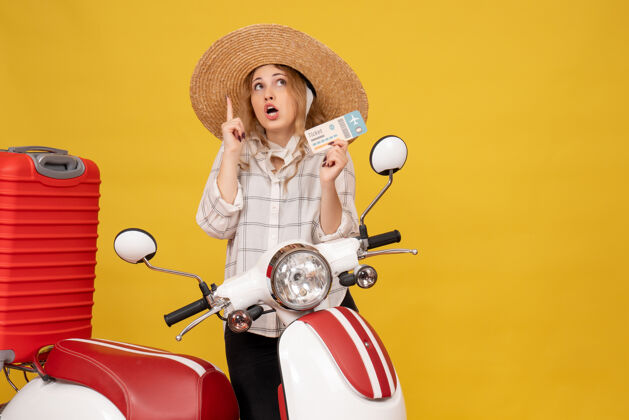 成人一个好奇的年轻女子戴着帽子坐在摩托车上拿着她的行李 把车票指着正前方坐着摩托车年轻女子