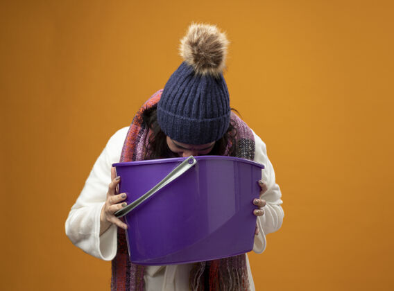 长袍年轻的生病的女人穿着长袍 戴着冬天的帽子和围巾有恶心拿着塑料桶呕吐到它隔离在橙色的墙上抱人年轻人