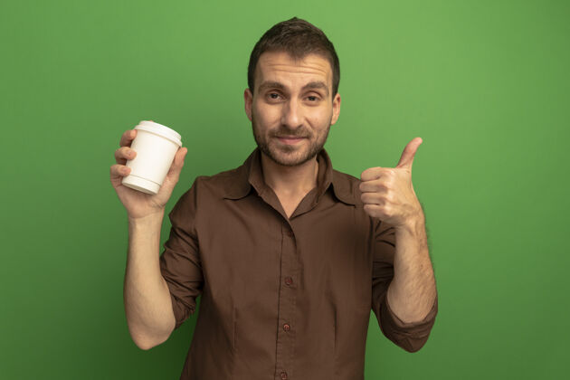 塑料高兴的年轻人看着前面拿着塑料咖啡杯 伸出大拇指孤立地站在绿色的墙上人人杯子