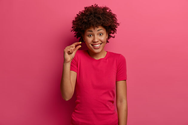 高兴积极的非裔美国女性展示一些非常小或很小的东西 在商店里讨论价格 开心地微笑 穿着随意 说一点点 孤立在粉红色的墙壁上尺寸概念表情人休闲
