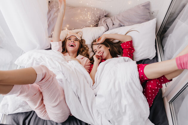 睡衣快乐的女模特躺在白毯子下笑着室内拍摄两个快乐的女孩在床上度过周末的早晨情绪微笑舒适
