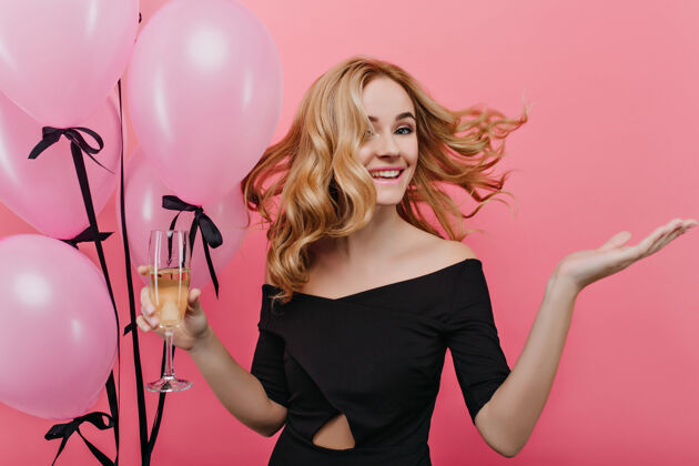 肖像情绪化的金发卷发女孩在生日派对上拿着酒杯跳舞穿着黑色服装的漂亮年轻女模特拿着粉色气球摆姿势舞蹈年轻优雅